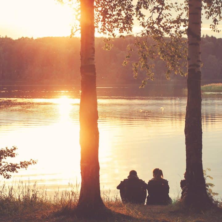 Couple sitting together between two trees at a lake at dusk having a big conversation.
