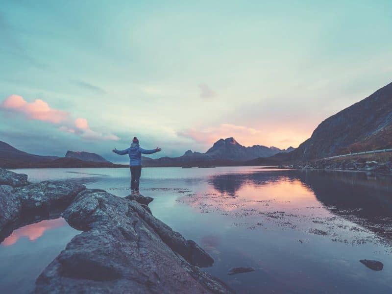 An adventerous woman standing on the edge of a rock with her arms up looking out at a lake and mountains at sunset.