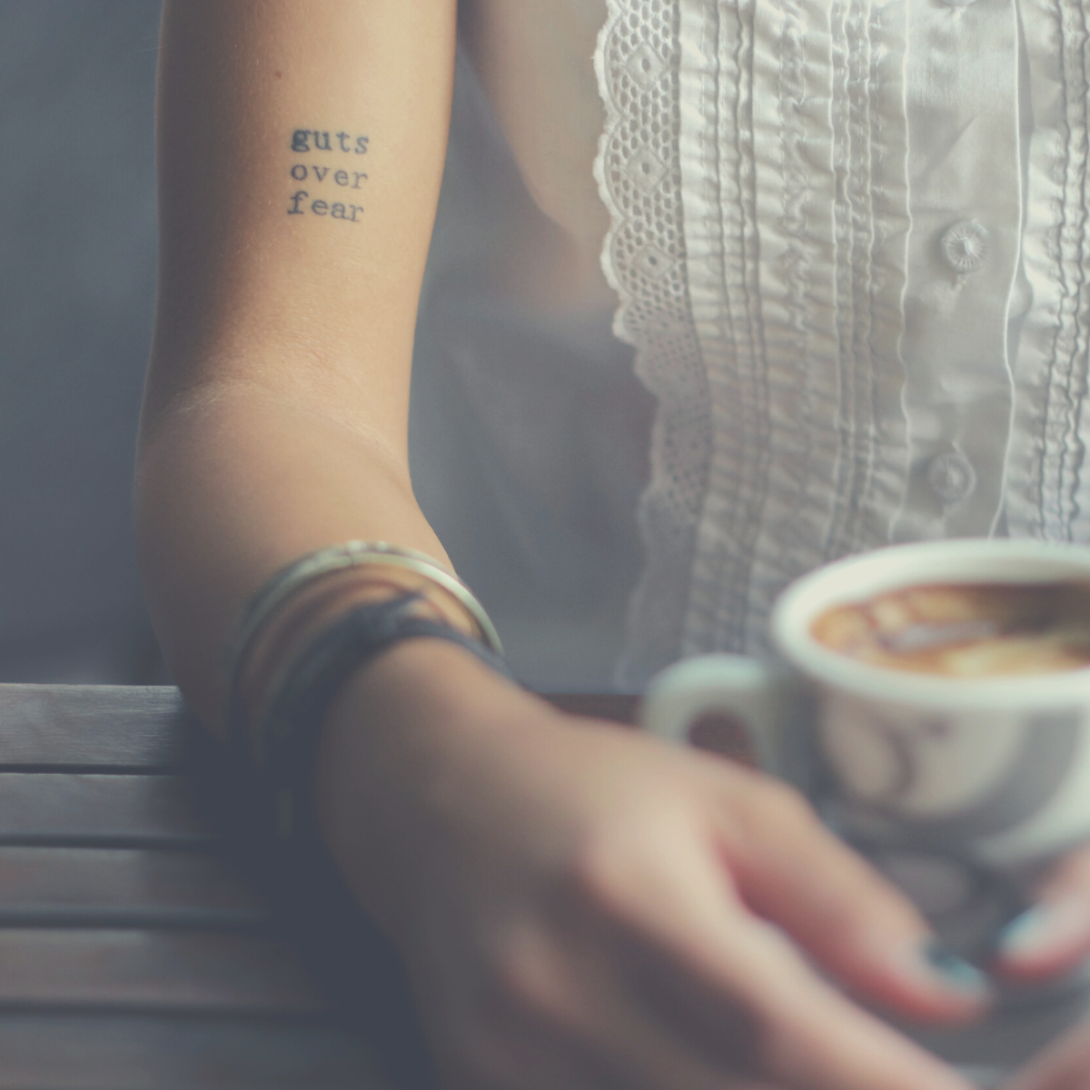 Woman with a cup of coffee and an arm tattoo that says, "guts over fear."