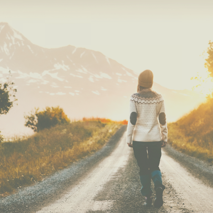 Fashionable woman walking alone down a road surrounded by grass and mountains.