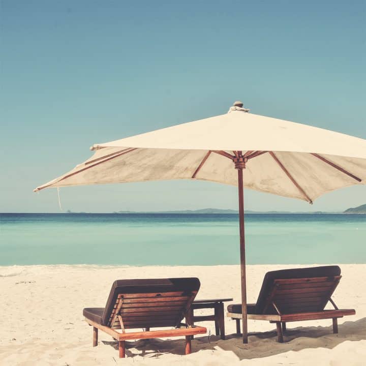 Two wood beach chairs sitting on white sand overlooking the ocean with a large canvas beach umbrella between them.