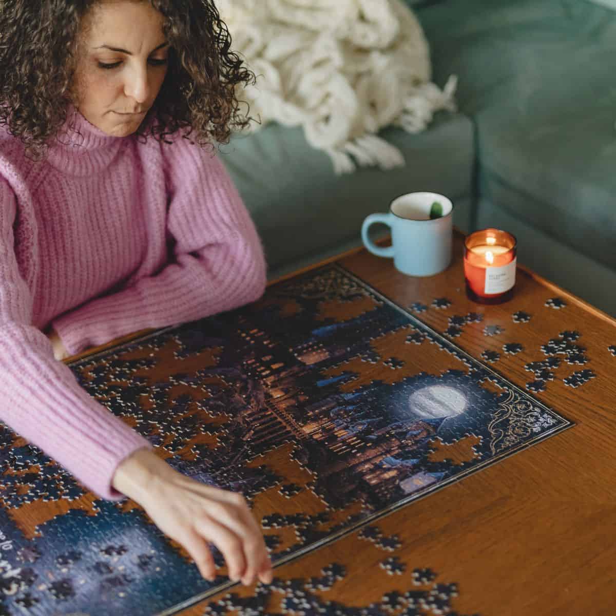 A woman sitting at a coffee table with a candle lit and a cup of tea working on a 1000 piece puzzle.