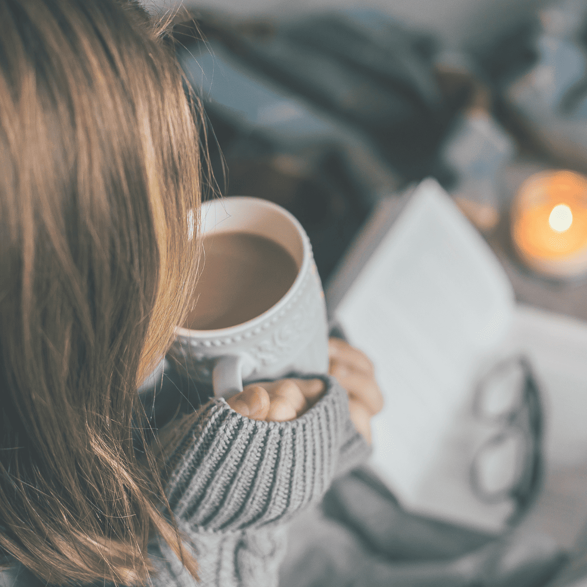 A woman sipping on a cup of hot cocoa practicing winter self-care and reading with candle.