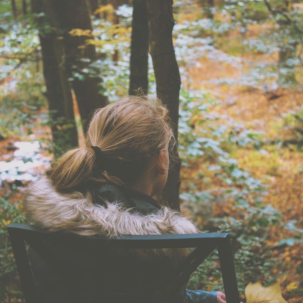 Woman struggling with Autumn Blues, sitting on a chair outside with a winter coat on, looking into the woods full of fall leaves.