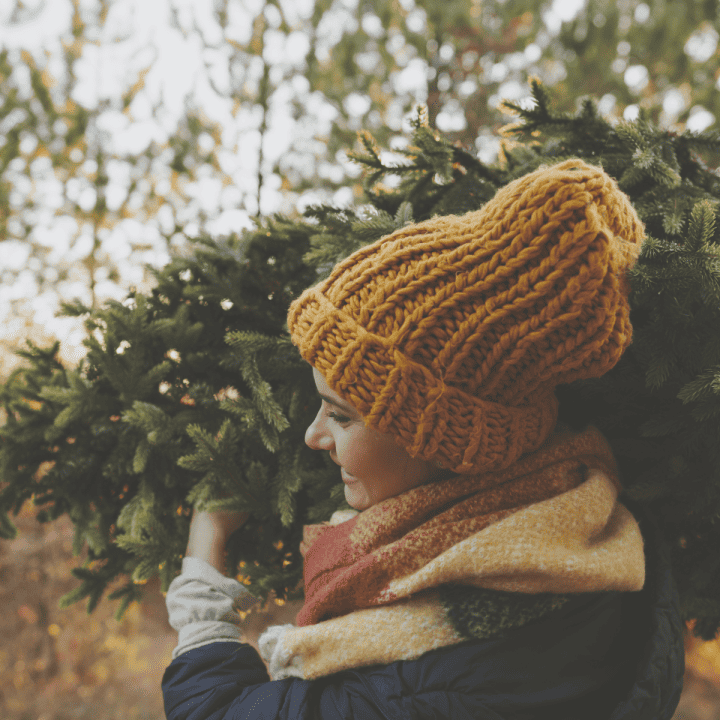 A happy woman wearing a hat and coat carrying a Christmas tree, slowing down and embracing the holiday season.