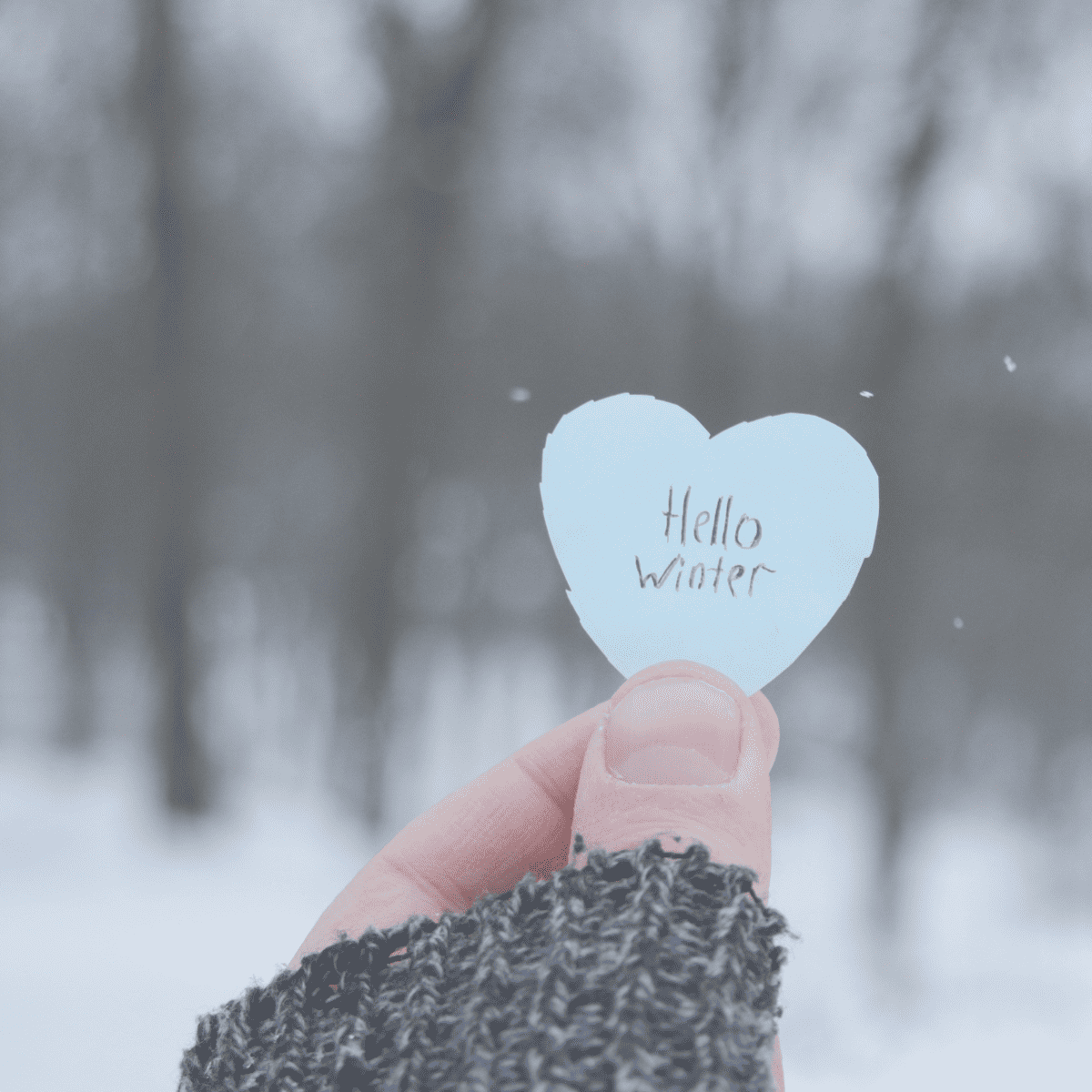 Woman holding a cut out paper heart with "hello winter" written on it.